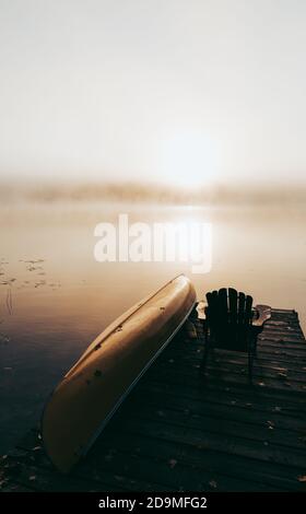 Leeres Dock mit umgedrehten Kanu an einem nebligen Morgen auf einem ruhigen See. Stockfoto