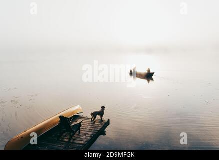 Zwei Personen im Fischerboot im Nebel mit Hund beobachten vom Dock aus. Stockfoto