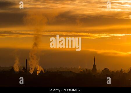 East Lothian, Schottland, Großbritannien, 6. November 2020. UK Wetter: Ein trüber Sonnenuntergang schafft einen nebligen Blick über die Skyline von Haddington mit dem Turm des Town House und den rauchenden Schornsteinen der Pure Malt Verarbeitungsfabrik sichtbar vor einem orangen Himmel silhouettiert Stockfoto