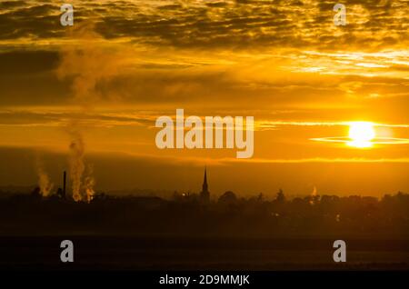 East Lothian, Schottland, Großbritannien, 6. November 2020. UK Wetter: Ein trüber Sonnenuntergang schafft einen nebligen Blick über die Skyline von Haddington mit dem Turm des Town House und den rauchenden Schornsteinen der Pure Malt Verarbeitungsfabrik sichtbar vor einem orangen Himmel silhouettiert Stockfoto