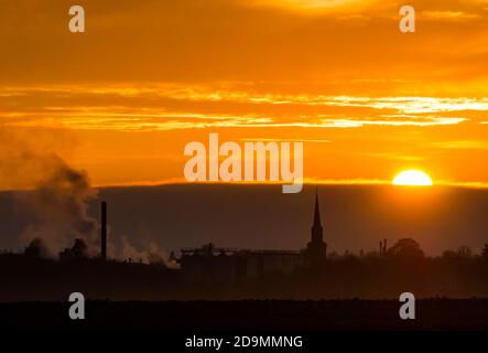 East Lothian, Schottland, Großbritannien, 6. November 2020. UK Wetter: Ein trüber Sonnenuntergang schafft einen nebligen Blick über die Skyline von Haddington mit dem Turm des Town House und den rauchenden Schornsteinen der Pure Malt Verarbeitungsfabrik sichtbar vor einem orangen Himmel silhouettiert Stockfoto