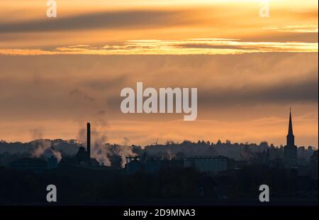 East Lothian, Schottland, Großbritannien, 6. November 2020. UK Wetter: Ein trüber Sonnenuntergang schafft einen nebligen Blick über die Skyline von Haddington mit dem Turm des Town House und den rauchenden Schornsteinen der Pure Malt Verarbeitungsfabrik sichtbar vor einem orangen Himmel silhouettiert Stockfoto