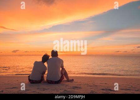 Ein Paar liebt es, den Sonnenuntergang an den Sommerferien am Strand zu beobachten. Die Silhouette der Menschen von hinten sitzend und genießen den Blick auf das Meer bei Sonnenuntergang auf tropischem Meer Stockfoto