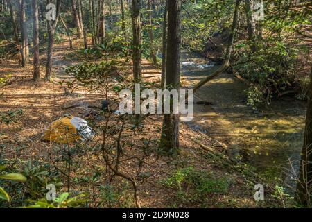 Blick vom Trail auf einen Mann, der in zelte Die Wälder entlang des Baches mit einem Zelt in der Schatten mit Rauch aus dem Lagerfeuer auf einem sonnigen Stockfoto