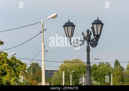 Retro-Straßenlampe mit schmiedeeisernen Elementen neben modernem urbanen Lichtmast. Konzept von Vergangenheit und Gegenwart, Vergleich, Technologieentwicklung. Stockfoto