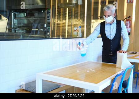 Kaukasischer Arbeiter Reinigungstisch mit Desinfektionsmittel in einem Restaurant während Coronavirus Ausbruch. Kellner in Schutzmaske Desinfektionstisch mit Stockfoto