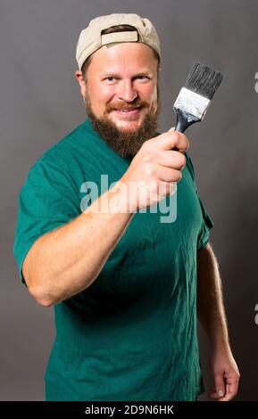 Fröhlicher bärtiger Vorarbeiter in grünem T-Shirt mit Pinsel Studio Portrait Auf grauem Hintergrund Stockfoto