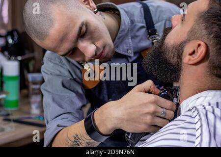 Bartstylen und Schneiden mit Trimmer. Junger bärtiger Hipster-Mann bekommt von einem professionellen Friseur in einem modernen Friseursalon einen Haarschnitt / Bart geschnitten. Stockfoto