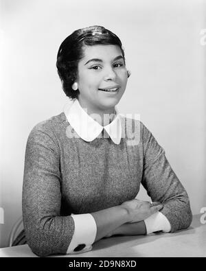 1950S 1960S PORTRAIT LÄCHELNDE AFROAMERIKANISCHE FRAU, DIE AUF DEN ARMEN GEFALTET LEHNT AUF DEM TISCH MIT BLICK AUF DIE KAMERA - N158 HAR001 HARS LADIES PERSONEN VERTRAUEN B&W AUGENKONTAKT GLÜCK FRÖHLICHE STILE AFRO-AMERIKANER AFROAMERIKANER SCHWARZ ETHNIZITÄT STOLZ LÄCHELT FRÖHLICH STILVOLLE MODE JUNGE ERWACHSENE FRAU SCHWARZ UND WEISS HAR001 ALTMODISCHE AFROAMERIKANER Stockfoto