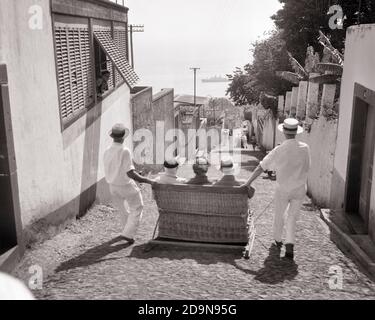 1950ER JAHRE ZWEI MÄNNER CARREIROS CHAUFFEURE FÜHREN DREI TOURISTEN IN EINEM RODELBAHN MONTE WICKER NACH FUNCHAL MADEIRA PORTUGAL - R15493 RGE001 HARS EUROPE B&W RODEL HOCHWINKEL ABENTEUER BERGAB EUROPÄISCHE INSELN TOURISTENATTRAKTION BESETZUNGEN SCHLITTEN STEIL ANONYM MADEIRA ATLANTIK OCEAN MONTE PORTUGAL FAHRGESCHÄFTE LÖSUNGEN SCHWARZ-WEISS STEIGUNG ALT MODISCH Stockfoto