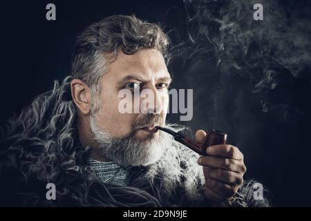 Solider bärtiger Mann in einem Pelzmantel rauchenden Pfeifenstudio Porträt auf schwarzem Hintergrund Stockfoto