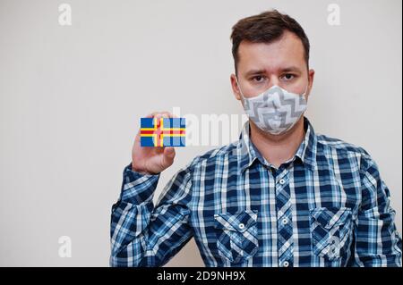 Mann im karierten Hemd zeigen Åland Flagge Karte in der Hand, tragen Schutzmaske isoliert auf weißem Hintergrund. Europa Länder Coronavirus Konzept. Stockfoto