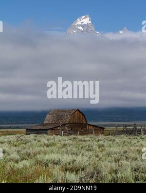 Die alte Holzscheune auf dem John Moulton Gehöft in der Mormon Row im Grand Teton National Park mit dem Grand Teton, der aus den Wolken dahinter guckt. Wyom Stockfoto