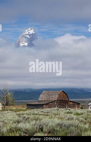 Die alte Holzscheune auf dem John Moulton Gehöft in der Mormon Row im Grand Teton National Park mit dem Grand Teton, der aus den Wolken dahinter guckt. Wyom Stockfoto