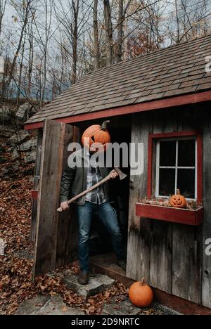 Mann trägt gruselig geschnitzten Kürbiskopf in Schuppen mit Axt für Halloween. Stockfoto