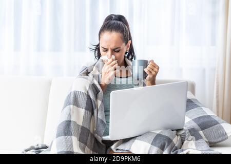 Home Office einer jungen Frau mit einer schweren Erkältung, die sich mit einer Decke bedeckt, niest und Tee trinkt. Stockfoto