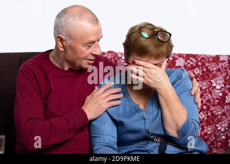 Ältere Ehepartner sitzen zu Hause auf der Couch, die Frau weint, der Mann umarmt sie und tröstet sie. Trauriges Lebensereignis Stockfoto