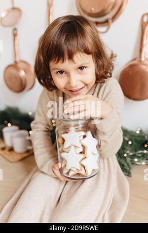 Kleinkind Mädchen tragen beige gestrickt Kleid hält Glas Glas mit weihnachtsplätzchen Stern Form. Minimalistisches Weihnachtskonzept Stockfoto