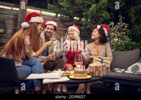 Eine Gruppe von Freunden toasten mit Weingläsern, feiern Neujahr, tragen santa Klausel Hüte, sitzen im Freien im Hinterhof Stockfoto