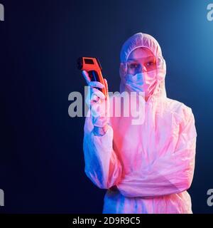 Krankenschwester in Maske und weiße Uniform steht in neon beleuchteten Raum und hält Infrarot-Thermometer. Stoppen Sie die Ausbreitung des Coronavirus Stockfoto