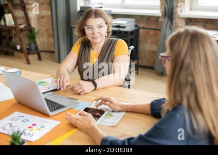 Junge lächelnde Geschäftsfrau in Casualwear, die Kollegen während der Diskussion ansieht Stockfoto