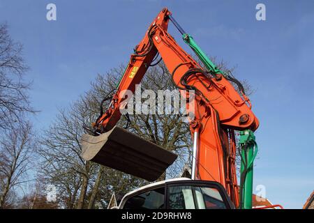 Orangefarbener hydraulischer Kolbenarm mit einer grauen Schaufel eines Baggers gegen einen blauen Himmel und blattlose Bäume in den Niederlanden im Herbst. November Stockfoto