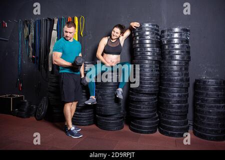 Schwangerschaftszeit bei sportlich starken muskulösen Frauen. Gemeinsame Workouts und Übungen des Paares während der Schwangerschaft in der Turnhalle. Mann und schwangere Frau Stockfoto