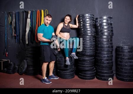 Schwangerschaftszeit bei sportlich starken muskulösen Frauen. Gemeinsame Workouts und Übungen des Paares während der Schwangerschaft in der Turnhalle. Mann und schwangere Frau Stockfoto