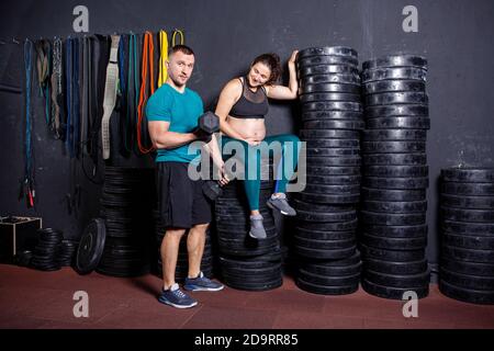 Schwangerschaftszeit bei sportlich starken muskulösen Frauen. Gemeinsame Workouts und Übungen des Paares während der Schwangerschaft in der Turnhalle. Mann und schwangere Frau Stockfoto