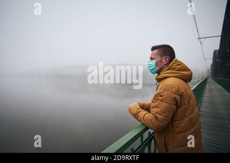 Einsamer Mann mit Gesichtsmaske auf Brücke gegen Stadt im Nebel. Themen Leben in neuer Normalität, Coronavirus und persönlicher Schutz. Stockfoto