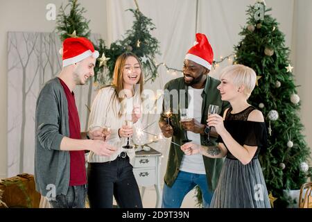 Frohe Männer und zufriedene Damen in Santa Hüte trinken Gläser Champagner während Kommunikation und Lachen auf Silvester Party. Sie halten bengalen Stockfoto