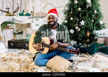 Schöner afroamerikanischer Junge in santa Hut hält giutar und bengalen Licht, sitzt auf dem Boden vor der Weihnachtsdekoration Stockfoto