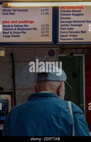 Ein älterer Herr, der eine Baseballmütze trägt und in einem Laden auf seine Fish and Chips wartet, wobei das Menü über seinem Kopf an der Wand steht. Stockfoto