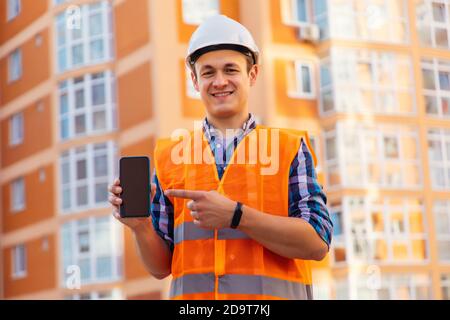 Architektur, Gebäude Business und Menschen Konzept. Lächelnder männlicher Architekt im Helm, der Telefon über dem Baustellenhintergrund zeigt Stockfoto