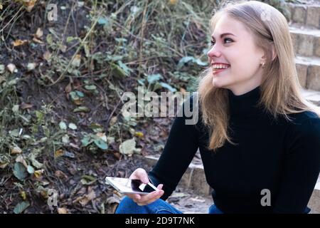 Junge blonde fröhliche Frau lachend sitzen mit Handy auf Ihre Hand draußen an einem herbstlich warmen Tag Stockfoto