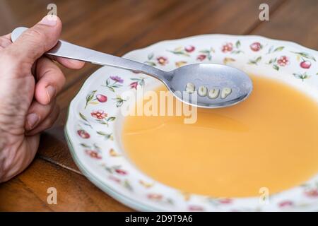 Eine Hand mit einem Löffel, der Alphabetsuppe in einer verzierten Schüssel auf einem Holztisch hält. Auf dem Löffel erscheint die Meldung Suppe Stockfoto