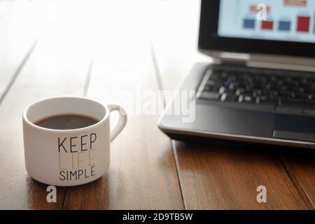 Kaffeetasse mit Nachricht halten das Leben einfach auf einem hölzernen Tisch und Computer im Hintergrund nicht fokussiert Stockfoto