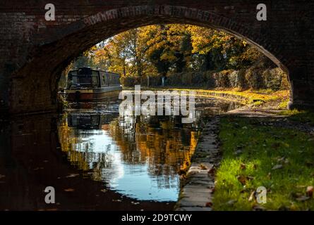 Ein Blick auf ein enges Boot unter dem Bogen einer gemauerten Fußbrücke, die sich in das stille Wasser des Kanals spiegelte, mit herbstlichen Farben in den Bäumen Stockfoto