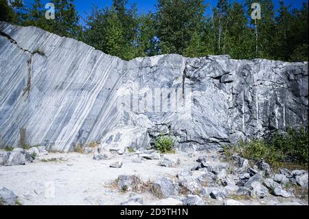 Felsenschnitt rohe Oberfläche aus weißem und grauem Marmor in der natürlichen Umgebung mit Bäumen und blauen Himmel. Blaue Marmor Karriere Textur. Hintergrund Stein, industrielles Material für Gebäude und Interieur. Stockfoto