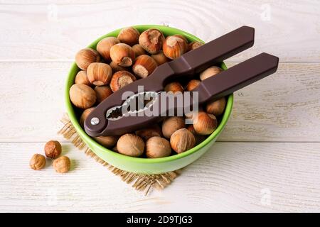 Brauner Nussknacker auf einer grünen Schüssel voller ungeschälter Haselnüsse über weißem Holztisch. Antioxidans und Proteinquelle für ketogene und rohe Ernährung. Stockfoto