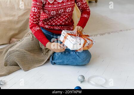Eine Frau wickelt ein Weihnachtsgeschenk, während sie zu Hause auf dem Boden sitzt. Bunte Bälle und Weihnachtsdekorationen auf dem Boden in der Nähe, Blick von oben. Stockfoto