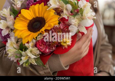 Die Person in der Lederjacke, die den schönen und bunten Blumenstrauß mit der Sonnenblume, den Rosen und verschiedenen Arten der Blumen, im Zimmer, dem Detail hält Stockfoto