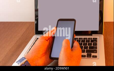 Hände eines Mannes, der ein Handy handhabt, das an einem Laptop mit einem leeren Bildschirm auf einem Holzschreibtisch mit warmer Beleuchtung befestigt ist, Mock up von Telefon und Laptop Stockfoto