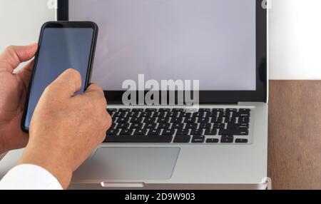 Hände eines Mannes, der ein Handy zusammenstellt Ein Laptop mit leerem Bildschirm auf einem Holzschreibtisch Stockfoto