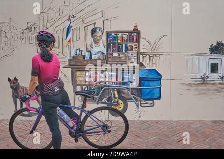 Eine Radfahrerin, die ein Wandgemälde mit einem Straßenhändler in der Ong Ang Walking Street von Klong Ong Ang, Chinatown / Phahurat, Bangkok, Thailand, betrachtet Stockfoto