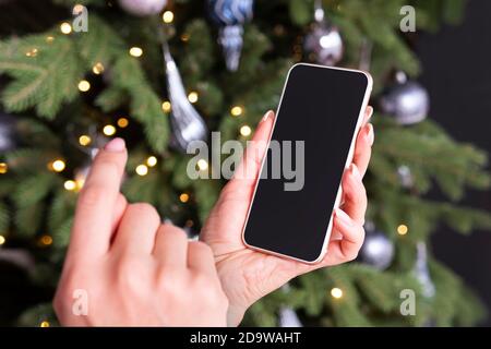 Smartphone in der Hand einer Frau auf dem Hintergrund eines Weihnachtsbaums. Leeren Raum auf dem Bildschirm für Design und Text. Das Mädchen tippt den Bildschirm mit ihrem f Stockfoto