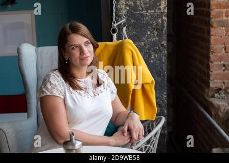 Schöne junge Frau mit roten langen Haaren in einer weißen Bluse, allein in einem Café auf einem Stuhl sitzend und auf ihre Bestellung wartend. Stockfoto