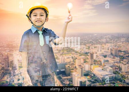 Doppelbelichtung des als Ingenieur gekleideten asiatischen Jungen Tragen einer Schutzkappe, die eine Glühlampe an einem hält Stadtlandschaft Kulisse-Phantasie und Kreativität c Stockfoto