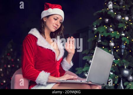 Eine junge Frau in einem Weihnachtsmann Kostüm sitzt vor einem Computerbildschirm und winkt mit den Händen. Weihnachtsgrüße online. Neujahr in Quarantäne. Stockfoto