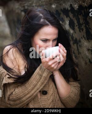 Natur, Schönheit, Jugend und gesundes Lifestyle-Konzept. Junge süße Mädchen in einem gestrickten Pullover vor einem Hintergrund einer alten Betonwand trinken Tee Fr. Stockfoto